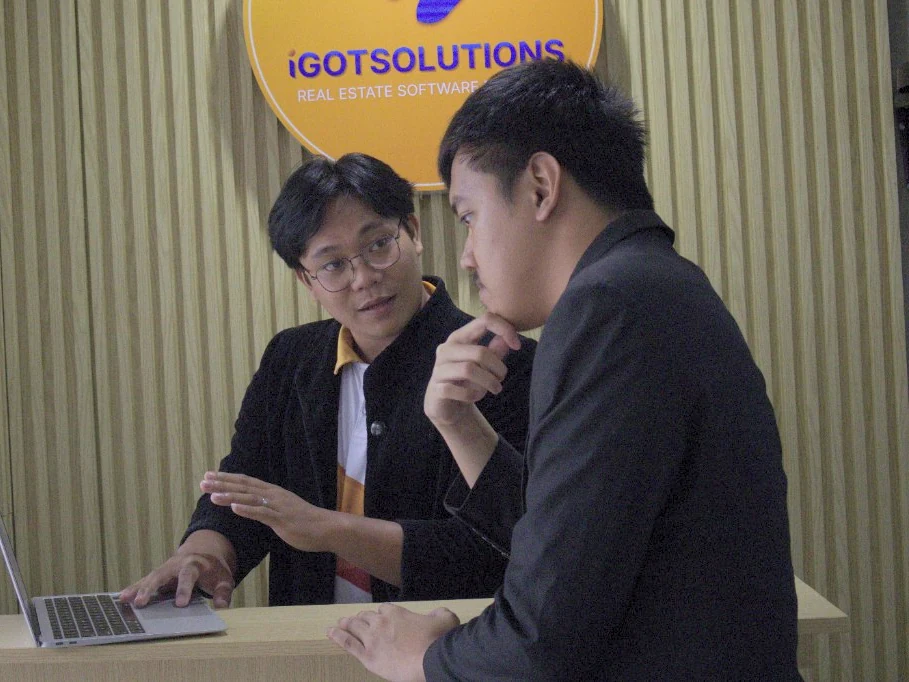Two iGotSolutions consultants reviewing a real estate software workflow on a laptop during a client session at the company's Cebu office, beneath the iGotSolutions Real Estate Software sign
